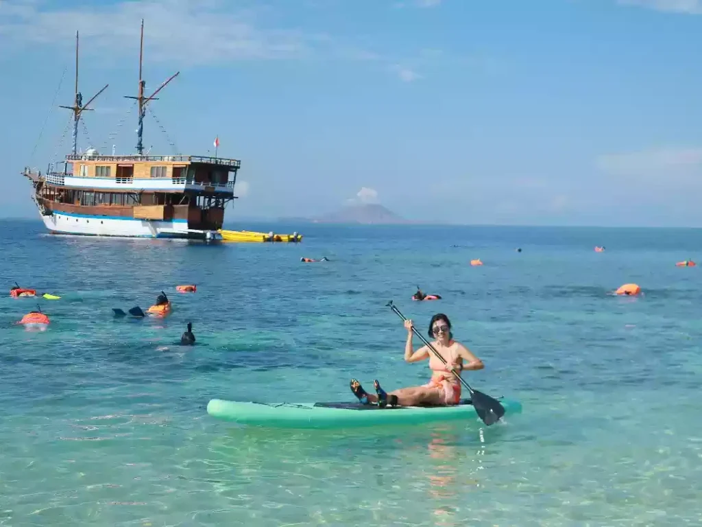 Guests paddleboarding beside an anchored phinisi in clear water